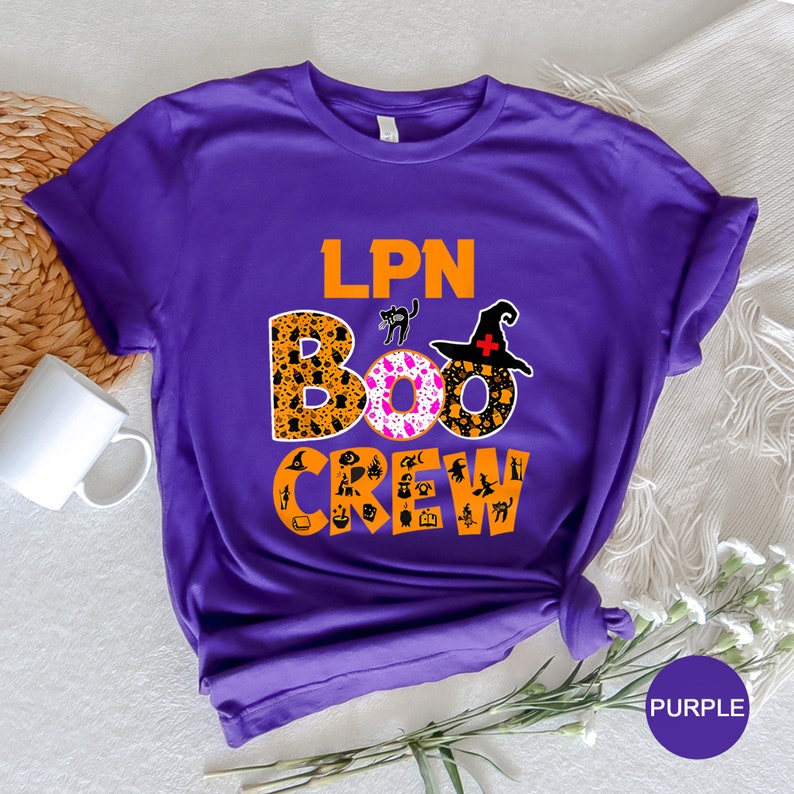 Pode incluir: Camiseta de manga curta roxa com o texto "LPN BOO CREW" em laranja e preto. O texto "BOO" &eacute; cercado por um padr&atilde;o rosa e laranja com &iacute;cones tem&aacute;ticos de Halloween.