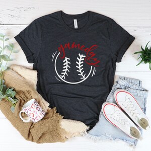 May include: Heather dark grey t-shirt with a white baseball graphic and the text "gameday" in red lettering.