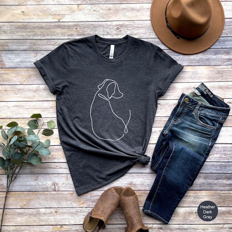 May include: A heather dark gray t-shirt with a white line drawing of a dog on the front. The shirt is shown on a wooden background with jeans, a hat, and shoes.