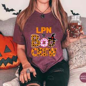 Pode incluir: Uma camiseta vinho com o texto "LPN BOO CREW" em preto e branco. O texto &eacute; cercado por gr&aacute;ficos tem&aacute;ticos de Halloween, incluindo um gato preto, um chap&eacute;u de bruxa e uma ab&oacute;bora.