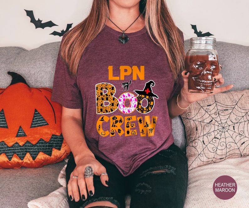 Pode incluir: Uma camiseta vinho com o texto "LPN BOO CREW" em preto e branco. O texto &eacute; cercado por gr&aacute;ficos tem&aacute;ticos de Halloween, incluindo um gato preto, um chap&eacute;u de bruxa e uma ab&oacute;bora.