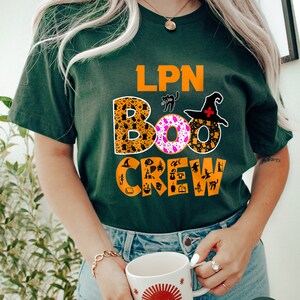 Pode incluir: Uma camiseta verde com o texto "LPN BOO CREW" em laranja e preto. As letras s&atilde;o decoradas com &iacute;cones tem&aacute;ticos de Halloween. A camiseta est&aacute; sendo usada por uma pessoa que est&aacute; segurando uma caneca com um design de sol vermelho.