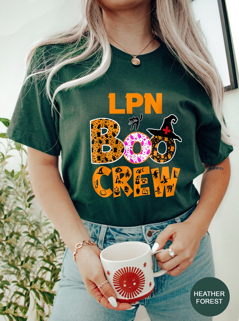 Pode incluir: Uma camiseta verde com o texto "LPN BOO CREW" em laranja e preto. As letras s&atilde;o decoradas com &iacute;cones tem&aacute;ticos de Halloween. A camiseta est&aacute; sendo usada por uma pessoa que est&aacute; segurando uma caneca com um design de sol vermelho.