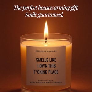 May include: A lit soy candle in a clear glass jar. The candle has a white label with the text "SMELLS LIKE I OWN THIS F*CKING PLACE". The text "The perfect housewarming gift. Smile guaranteed." is at the top.