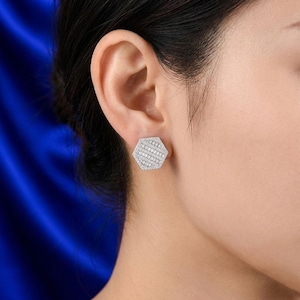 May include: A hexagonal stud earring featuring a silver-toned metal setting encrusted with numerous small, clear, rectangular baguette-cut stones arranged in a geometric pattern, creating a reflective and textured surface on the earlobe.