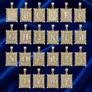 Op de afbeelding: Een collectie rechthoekige goudkleurige hangers, elk met een enkele zilverkleurige letter van A tot Y gecentreerd op een getextureerde gouden achtergrond. Elke hanger heeft een decoratieve geschulpte rand en een oogje aan de bovenkant.