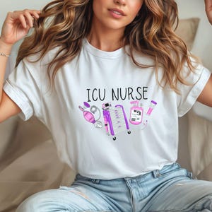 May include: White t-shirt with the text "ICU NURSE" in black letters. The shirt features illustrations of medical equipment in pink and purple, including a ventilator, a medical cart, and a bottle of hand sanitizer.