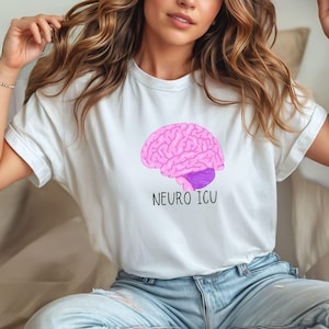 May include: White t-shirt featuring a pink brain graphic with a purple stem and the text "NEURO ICU" in black. The shirt is made of a soft material and is suitable for casual wear. The design is a medical or science-themed graphic.