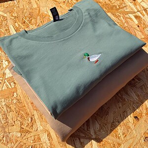 May include: A folded green t-shirt with a small embroidered image of a duck on the front. The t-shirt is folded on top of a brown t-shirt.