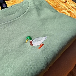 May include: A sage green t-shirt with an embroidered mallard duck in the center. The duck is white with a green head and orange feet.