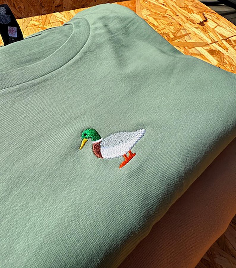May include: A sage green t-shirt with an embroidered mallard duck in the center. The duck is white with a green head and orange feet.