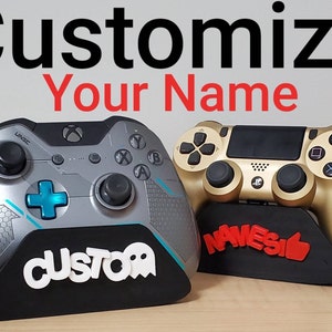 Könnte beinhalten: Zwei Videospielcontroller, einer silber und einer gold, werden auf kundenspezifischen Ständern präsentiert. Der silberne Controller ist ein Xbox-Controller und der goldene Controller ist ein Playstation-Controller. Die Ständer haben die Wörter "CUSTOM" und "NAMESI" aufgedruckt.