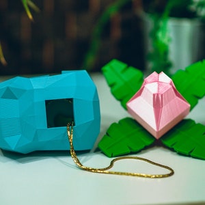 May include: A turquoise geometric box with a gold chain extending from a square opening. Beside it is a pink geometric flower with green leaves. The objects are on a white surface with green plants in the background.