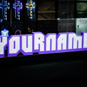 Peut inclure: Une plaque de nom en 3D imprimée en violet avec des lettres blanches qui épellent "YOURNAME".