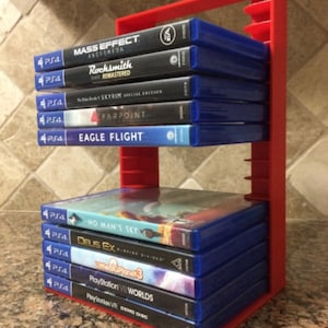 May include: A red plastic stand holds a stack of video game cases. The cases are blue and feature titles like "Mass Effect: Andromeda", "Rocksmith Remastered", "Skyrim Special Edition", "Farpoint", "Eagle Flight", "No Man's Sky", "Deus Ex: Mankind Divided", "LittleBigPlanet 3", "PlayStation VR Worlds", and "PlayStation VR Demo Disc".