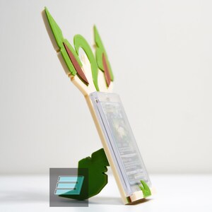 May include: A green and white 3D printed card holder shaped like a Pokemon character. The holder has a slot for a trading card and a base to stand on.