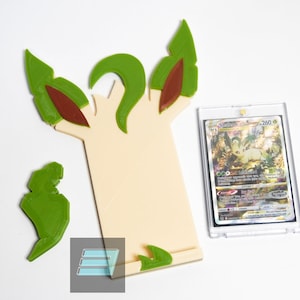 May include: A 3D printed card holder shaped like a tree with green leaves and brown branches. The holder is designed to hold a standard trading card, which is shown in the image. The card is a Pokemon card featuring the character Gardevoir.