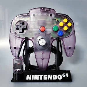 Op de afbeelding: Een transparante paarse Nintendo 64-controller met een zwarte standaard. De controller heeft een transparante paarse behuizing met zichtbare interne componenten. De standaard heeft de tekst "NINTENDO 64" op de voorkant.