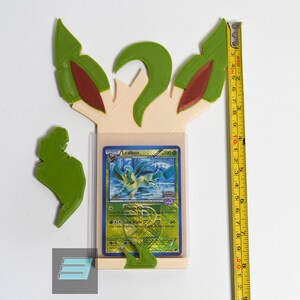 May include: A 3D printed stand for a Pokemon trading card, featuring a green and brown Leafeon design. The stand is made of a light beige plastic and has a slot for a standard-sized trading card. The card in the stand is a Leafeon card from the Pokemon Trading Card Game.