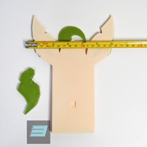 May include: A 3D printed model of a green and beige character with large ears. The model is approximately 10 inches tall and has a small hole in the center. The model is sitting on a white surface.