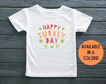 Puede incluir: Camiseta blanca con el texto "Happy Turkey Day" en letras naranja y rojas con hojas verdes alrededor del texto. El texto "AVAILABLE IN 6 COLORS!" está en un círculo naranja en la esquina inferior derecha de la imagen.