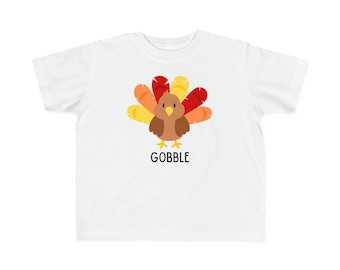 Puede incluir: Camiseta blanca para niño pequeño con una ilustración de pavo de dibujos animados y la palabra "GOBBLE" impresa debajo. El pavo tiene plumas marrones, plumas de cola rojas y naranjas y un pico amarillo.