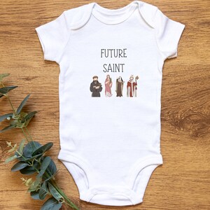 May include: White baby onesie with the text "FUTURE SAINT" printed in black. Four small images of saints are printed below the text.