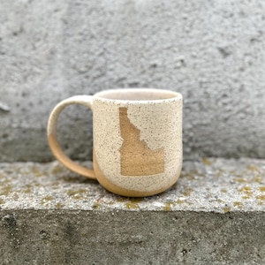 Può includere: Una tazza in ceramica bianca con una glassa macchiettata e un contorno dorato dello stato dell'Idaho. La tazza ha un manico ed è appoggiata su una superficie di cemento grigia.