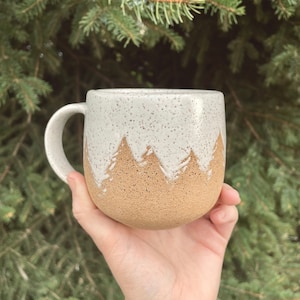 May include: A white ceramic mug with a speckled glaze and a brown mountain design. The mug has a rounded shape and a handle.