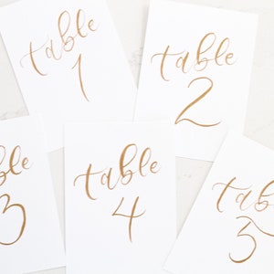 May include: Five white cards with gold calligraphy that read "table" followed by a number from one to five. The cards are arranged at different angles, with the numbers in a handwritten script.
