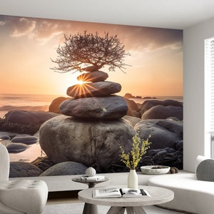 May include: A stack of gray stones balanced on top of each other with a small tree growing out of the top stone. The stones are on a beach with the ocean in the background. The sun is setting in the distance, creating a warm glow over the scene.