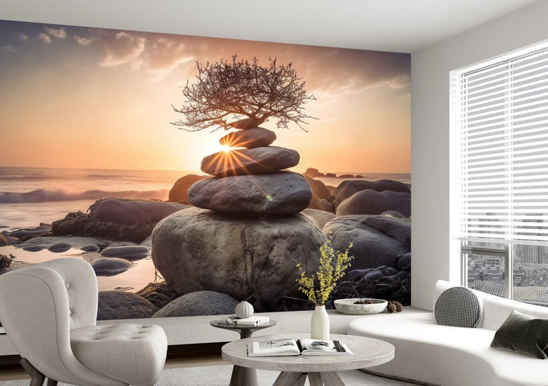 May include: A stack of gray stones balanced on top of each other with a small tree growing out of the top stone. The stones are on a beach with the ocean in the background. The sun is setting in the distance, creating a warm glow over the scene.