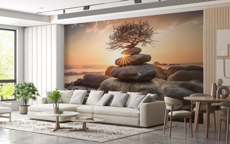 May include: A stack of gray stones balanced on top of each other with a small tree growing out of the top stone. The stones are on a beach with a sunset in the background.