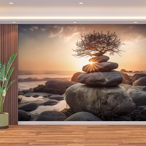 May include: A stack of gray stones balanced on top of each other with a small, bare tree growing out of the top stone. The stones are on a rocky beach with a sunset in the background.