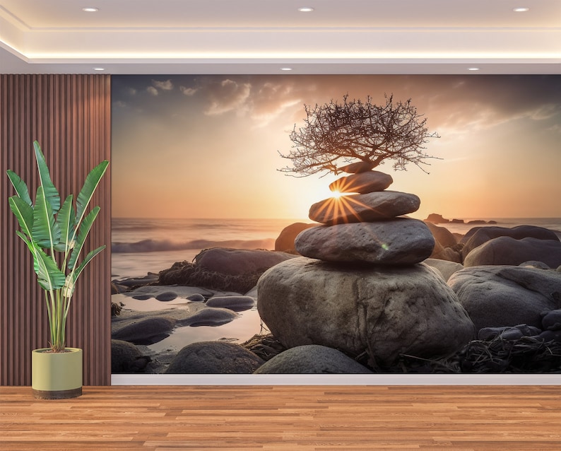 May include: A stack of gray stones balanced on top of each other with a small, bare tree growing out of the top stone. The stones are on a rocky beach with a sunset in the background.