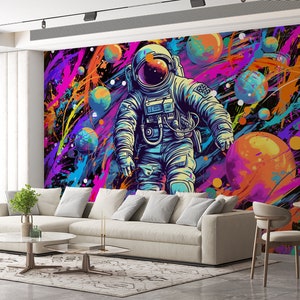 May include: An astronaut in a blue and white spacesuit floats in a colorful galaxy with planets and stars. The background is a dark blue with splashes of pink, purple, yellow, and orange paint.