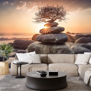May include: A stack of gray stones balanced on top of each other with a small tree growing out of the top stone. The stones are on a beach with a sunset in the background.
