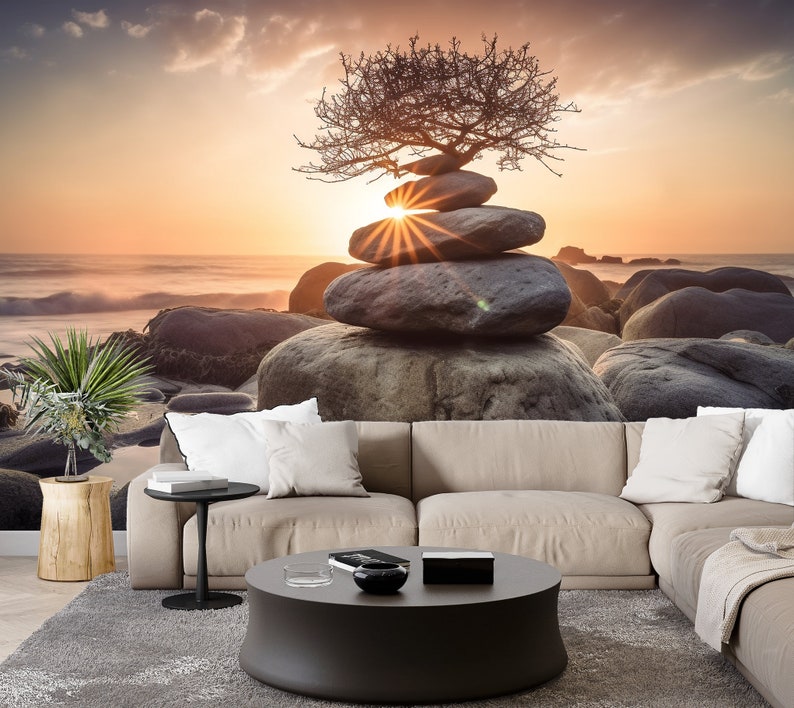 May include: A stack of gray stones balanced on top of each other with a small tree growing out of the top stone. The stones are on a beach with a sunset in the background.
