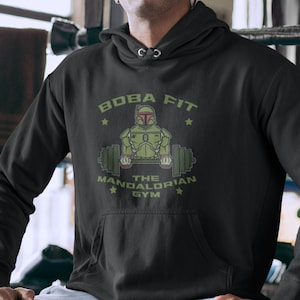May include: Black hooded sweatshirt with a green and red graphic. The graphic features a cartoon image of a Mandalorian character lifting weights, with the text "BOBA FIT THE MANDALORIAN GYM".