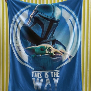 May include: Blue fleece blanket with a white circle design featuring a Mandalorian and Grogu. The text "This is the Way" is printed in white below the circle.
