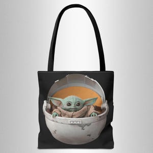 Puede incluir: Una bolsa de mano negra con una imagen blanca y gris de un personaje de la franquicia Star Wars, The Mandalorian, conocido como "The Child" o "Baby Yoda", sentado en una c&aacute;psula flotante.
