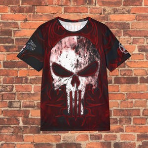 May include: Black t-shirt featuring a large, distressed white skull with black eye sockets and a vertical line design. The skull is set against a red background with tribal-style accents. The shirt has short sleeves and a crew neck.