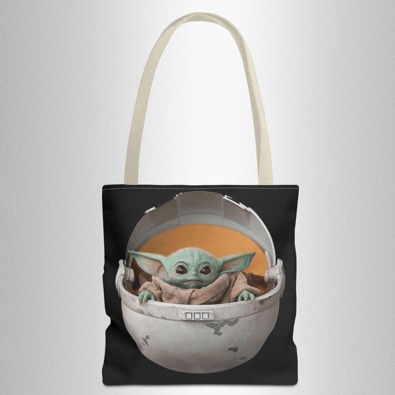 Puede incluir: Una bolsa de tela negra con un asa blanca presenta un gr&aacute;fico de un personaje alien&iacute;gena verde en una c&aacute;psula blanca con un interior naranja. El alien&iacute;gena tiene orejas grandes y est&aacute; mirando al espectador.