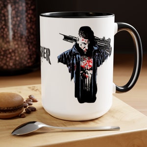 May include: White ceramic mug with a black rim and handle. The mug features a graphic of a man with a skull on his chest, holding a gun. The text "Punisher" is printed on the mug.