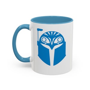 May include: White ceramic mug with a blue rim and a blue graphic of a stylised Mandalorian helmet on the front.