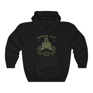 May include: Black hoodie with a green and tan graphic. The graphic shows a character in a helmet lifting weights, with the text "BOBA FIT" above and "THE MANDALORIAN GYM" below. The design includes stars and a barbell.