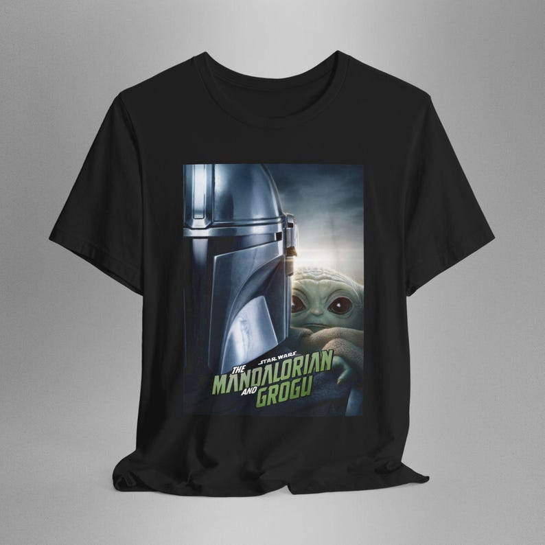 May include: Black t-shirt featuring a graphic from The Mandalorian series. The design includes the Mandalorian's helmet and Grogu, with the text "The Mandalorian and Grogu" in green and white.