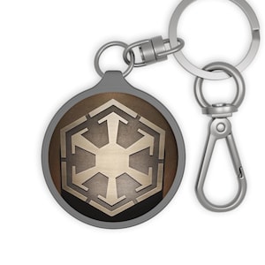 May include: A round keychain with a silver metal ring and clasp. The keychain features a brown and gold emblem with a geometric design. The emblem is set in a grey border.