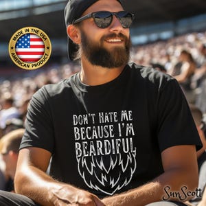 May include: A black t-shirt with a white graphic that says "Don't hate me because I'm beardful" with a stylized beard graphic below the text. The shirt is made in the USA.