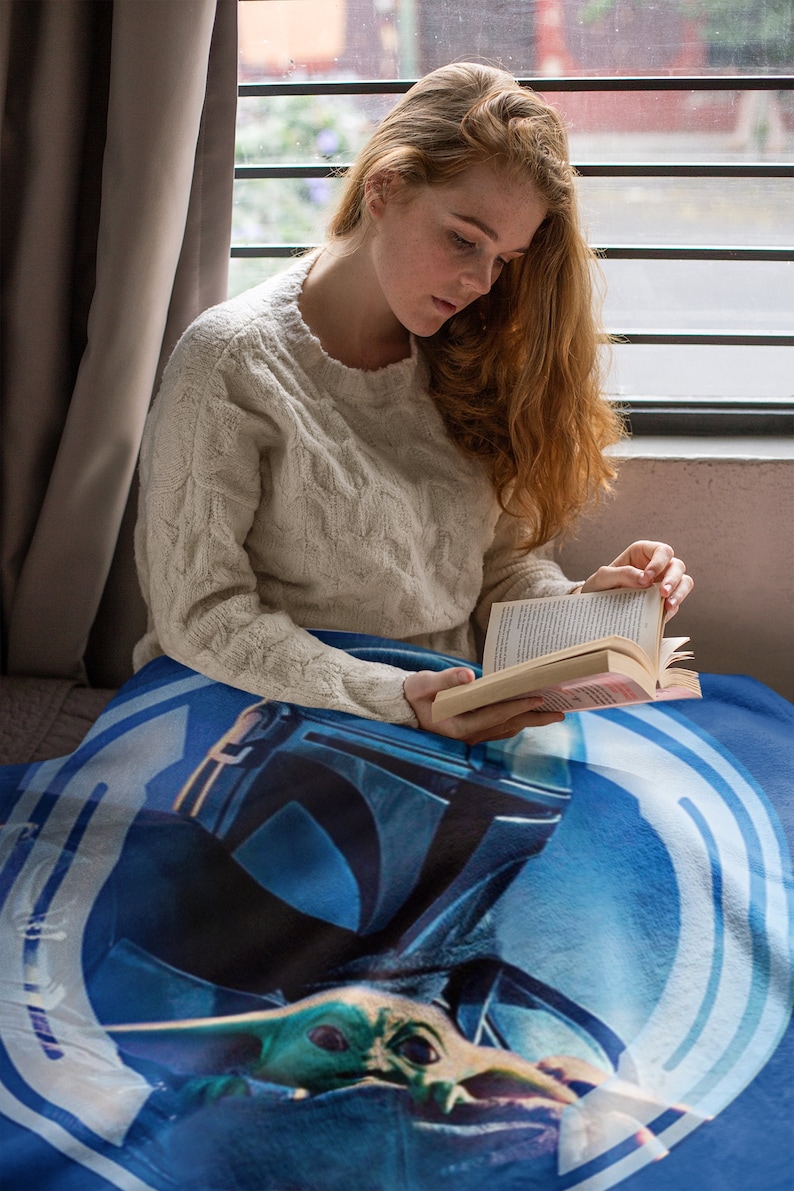 May include: A woman sits on a blue and white blanket with a Star Wars Mandalorian and Grogu design. She is reading a book and wearing a white sweater.